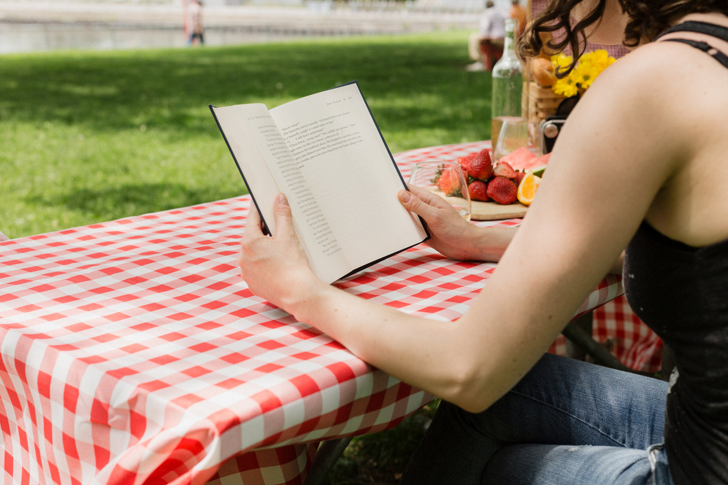 shoulders and arms of a woman readying a book at picnic table with a a red and white checkered tablecloth. 